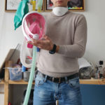 A light-skinned man carries a transparent snorkel mask with a pink interior, white frame and two yellow valves on top, each attached to long ribbed tubes, a turquoise air bag connects to one valve.