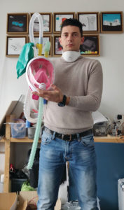 A light-skinned man carries a transparent snorkel mask with a pink interior, white frame and two yellow valves on top, each attached to long ribbed tubes, a turquoise air bag connects to one valve.