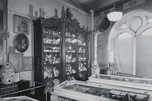 A black-and-white photograph of a museum gallery, with a large, ornate cabinet holding ceramics, paintings on the wall, and an arched window.