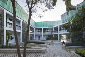 Interior courtyard of GHESKIO hospital, a two-story, angular white building with a luminescent green sloping roof that has large rectangular windows and a wrap-around balcony. The cobbled courtyard has climbing green foliage in triangular stone planters.