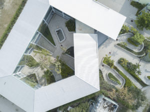 Aerial view of GHESKIO hospital, the building is shaped like a capital letter G that has slanted, ridged white roofs with a central paved courtyard split by triangular and oblong planters