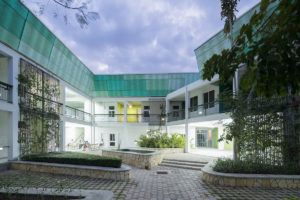 Interior courtyard of GHESKIO hospital, a two-story, angular white building with a luminescent green sloping roof that has large rectangular windows and a wrap-around balcony. The cobbled courtyard has climbing green foliage in triangular stone planters.