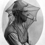 Photograph of a man sitting while wearing a mosquito net veil with a smoking pipe in his mouth