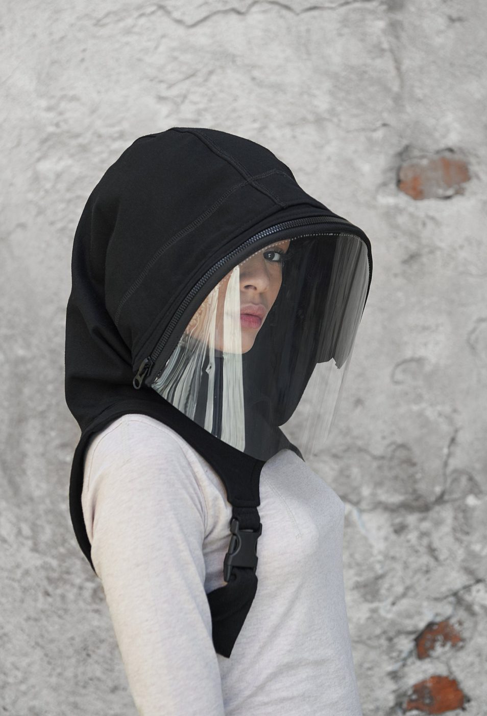 Woman wearing black hood that extends to her upper back and is secured under her arms with buckling straps. A zipper connects the front of the hood to a clear face shield.