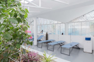 On a gray veranda under a white pitched roof are several blue cots similar in size to beach chairs that can be angled up for patients to recline on them; at the center of each cot is a hole.