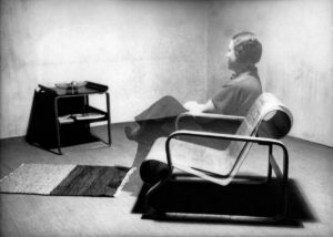 Photo of a woman's translucent form sitting in a curved plywood chair looking toward the wall.