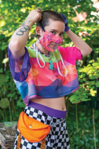 Photograph of a light-skinned individual dressed in bright colors adjusting a pink and purple face mask with a clear mouth panel, standing against a sun-dappled leafy green backdrop.