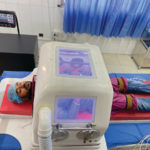 A medium-skinned figure lies with eyes closed on a hospital bed underneath a box-shaped negative pressure air ventilator which is opaque with rectangular transparent planes revealing the chest and side