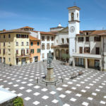 A plaza with numerous white squares painted on the ground one-point-eight meters apart in a lattice design for social distancing. The square is surrounded by buildings and has a statue in the center