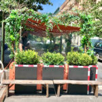 Side-view of an urban outdoor dining structure with several people dining behind plexiglass; it has black planters with green shrubs, wooden benches and a rust-colored awning overgrown with green ivy