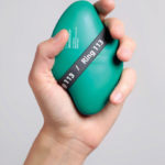 A light-skinned left hand holds a curved lozenge-shaped green-blue plastic device containing naloxone in a loose grip. The device is about the size of their palm and has text on the surface