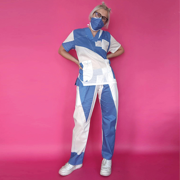 A woman wears blue and white hospital scrubs and face mask, set against a bright pink backdrop.