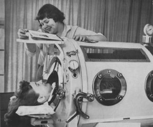 Black-and-white photograph showing a smiling woman attending to a young patient whose head protrudes from a large cylindrical iron lung with porthole-shaped openings along its side