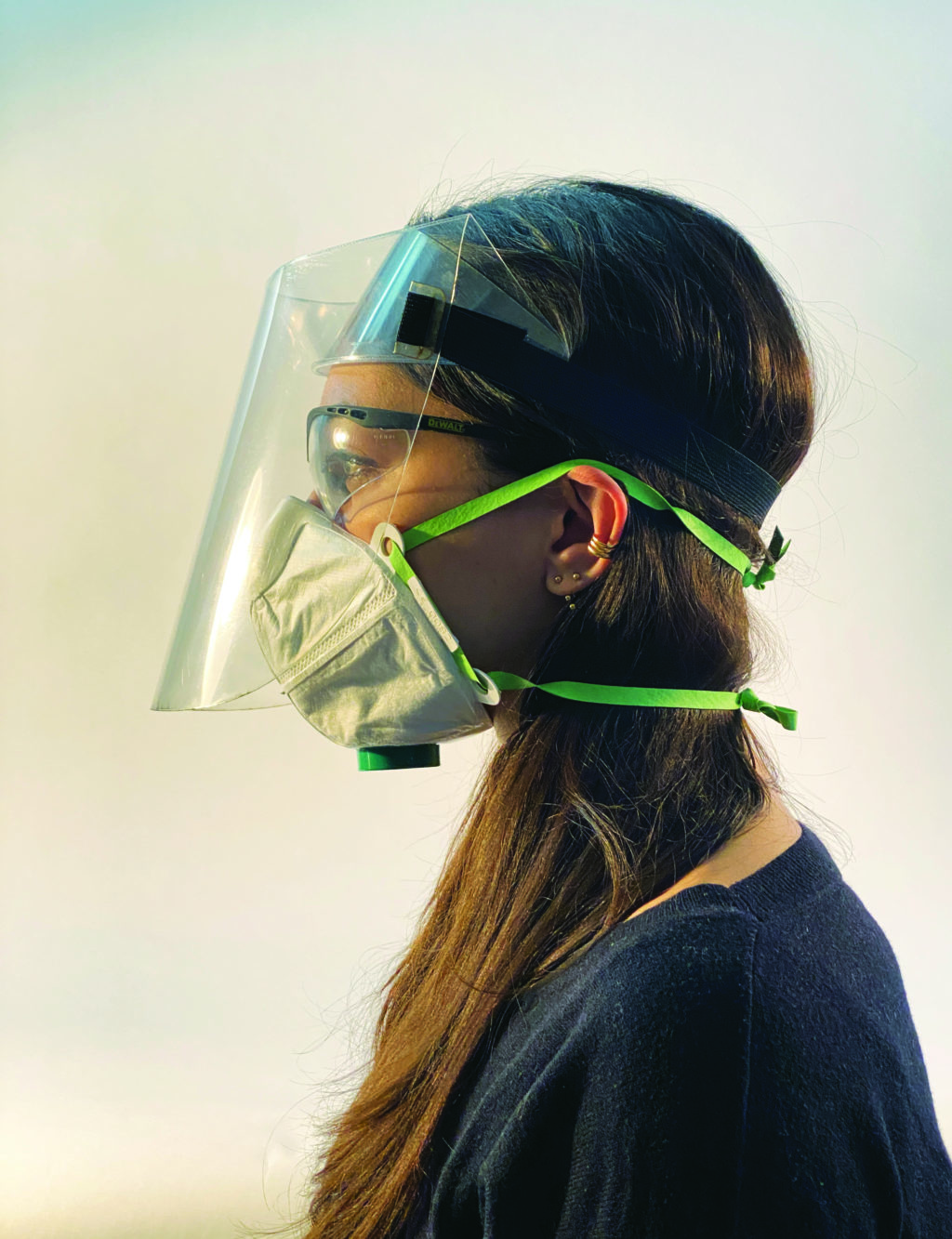 A woman in profile wearing a white face mask, safety glasses, and a folded clear face shield with a black elastic headband