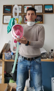 A light-skinned man holds a transparent snorkel mask with a pink interior, white frame and two yellow valves on top, each attached to long ribbed tubes, a turquoise air bag connects to one valve.
