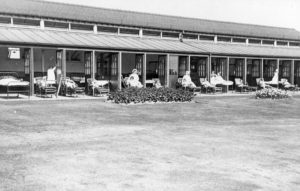 Black and white photograph of a two-story sanatorium with thirteen patient bedrooms opening out onto a lawn. The rooms have either beds or reclining chairs with patients sitting or standing beside them.