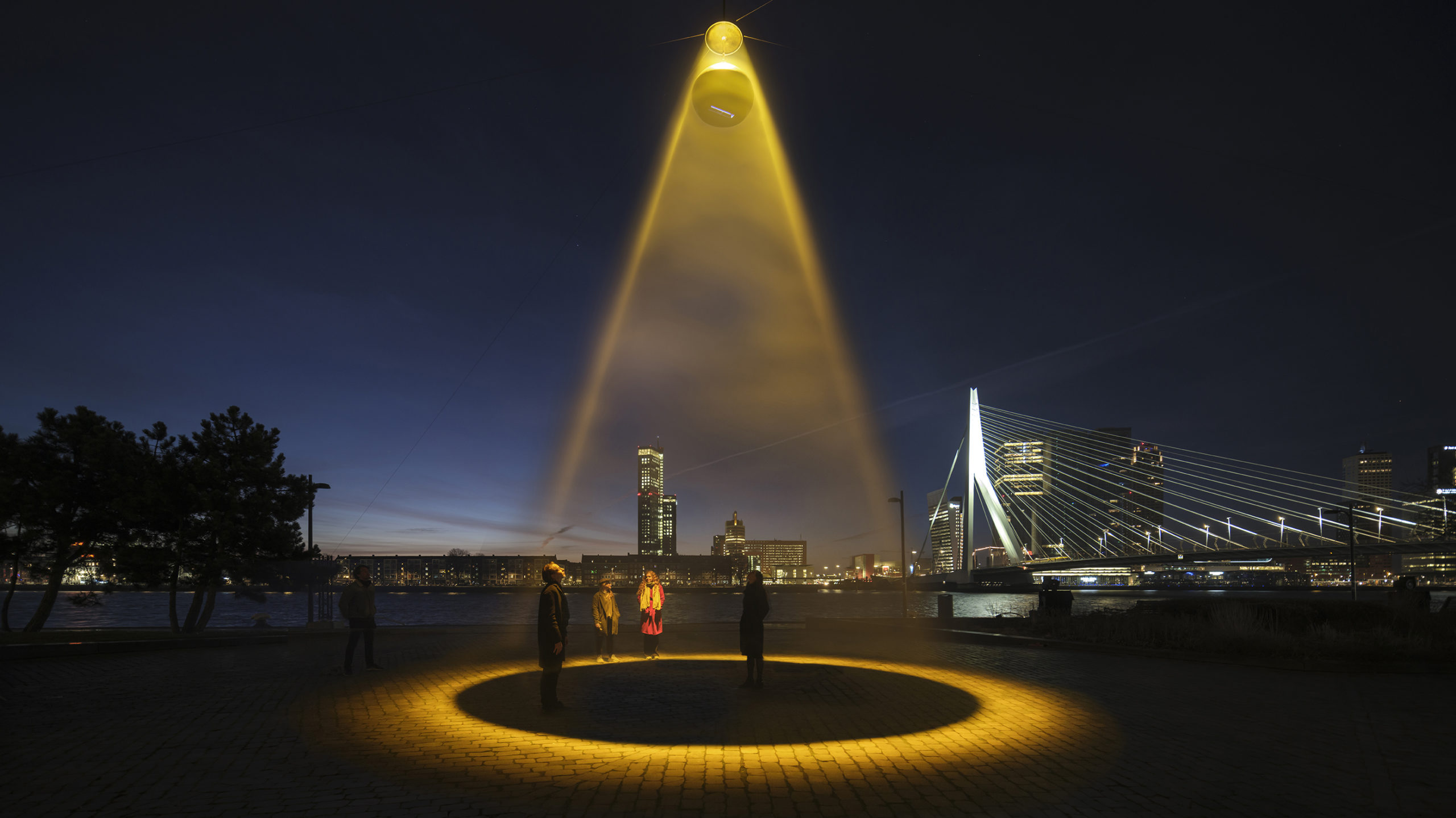A night-time photograph of a public space with a high suspended light source which illuminates a large yellowish open circle on the paved surface below.