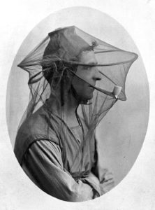 Photograph of a man sitting while wearing a mosquito net veil with a smoking pipe in his mouth