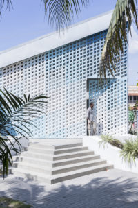 A light blue trapezoidal building with pointed roofline and angled stairs, its facade covered in a perforated panel with square holes of various sizes checkered in several shades of blue