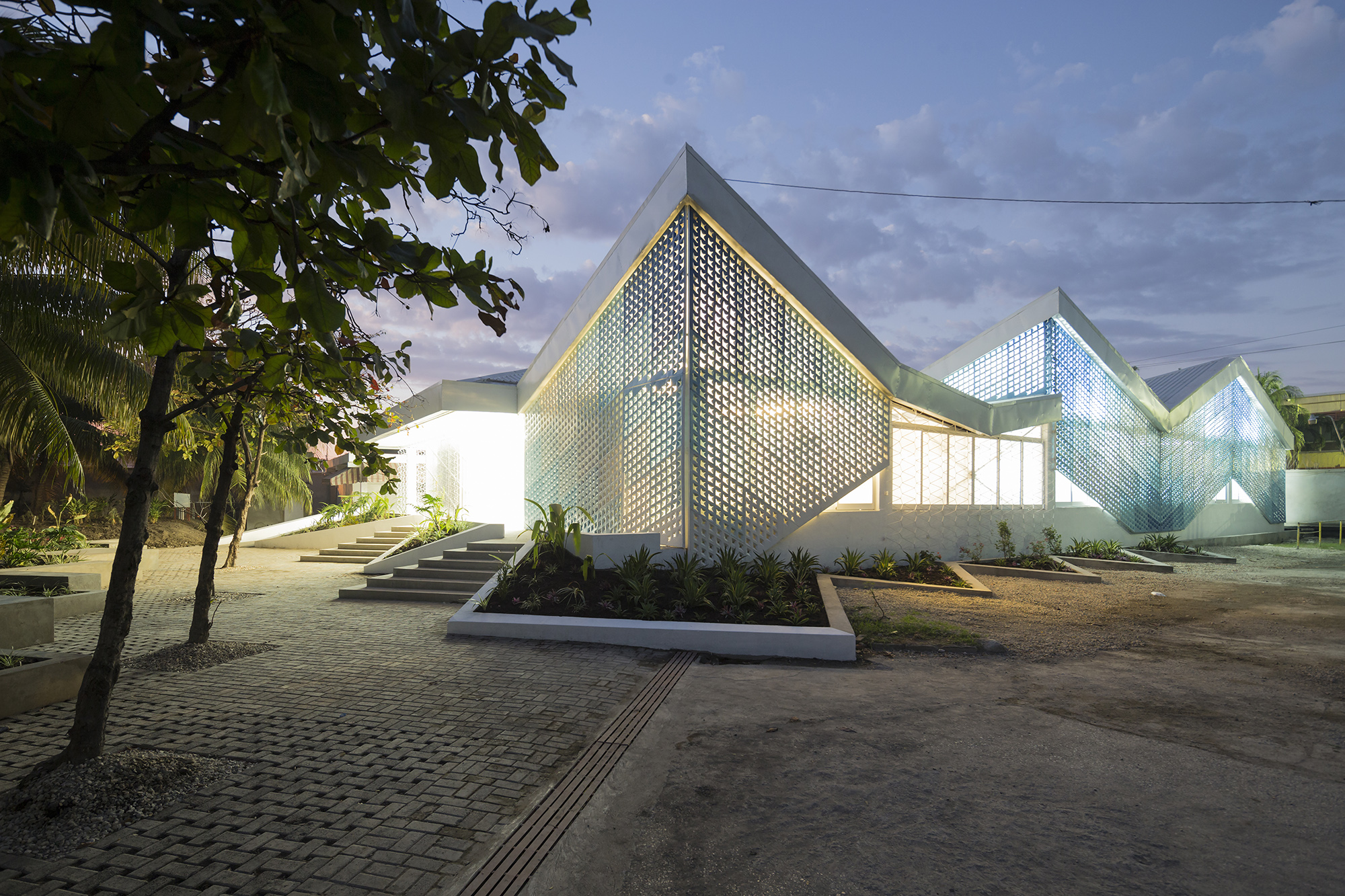 A glowing angled building with zigzagging roofline at dusk; light bleeds through perforated panels on the facade