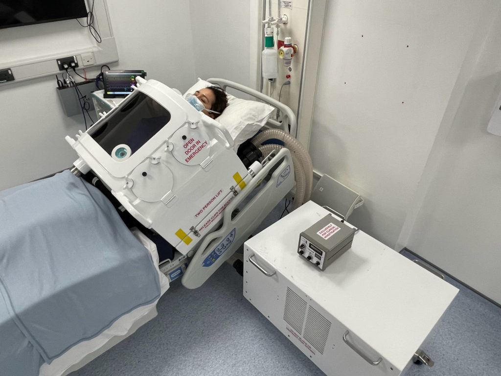 A patient wearing a face mask reclines on a hospital bed, their torso enclosed in a cylindrical ventilator with a large clear window at the front