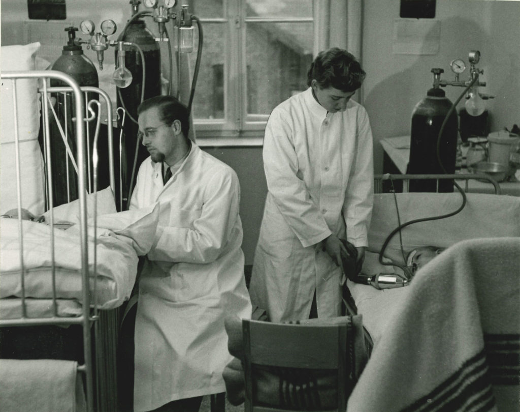 Black-and-white photograph depicting two medical professionals in white lab coats attending to patients in beds breathing through tubes and connected to large oxygen tanks