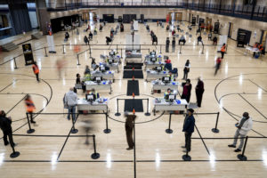 Inside a school gymnasium, health care workers sit at spaced tables to vaccinate individuals, who stand several feet from each other in lines marked by stanchions