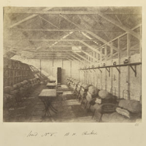Sepia photograph showing building interior with exposed rafters and rows of metal cots stacked with blankets and pillows