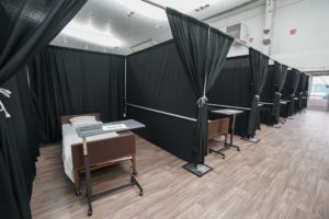 Inside an industrial interior, a view of makeshift patient rooms enclosed by black curtains, each with a small patient bed and rolling table