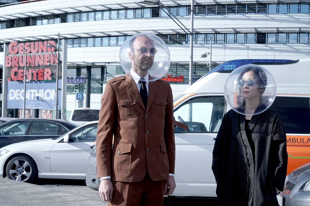 Two light-skinned figures wear large clear-plastic spheres over their heads and round sunglasses; one, on the left, wears a brown suit, the other wears a black coat, they stand in front of cars and a shopping center