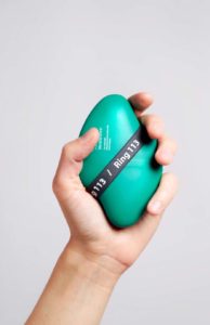 A light-skinned left hand holds a curved lozenge-shaped green-blue plastic device containing naloxone in a loose grip. The device is about the size of their palm and has text on the surface