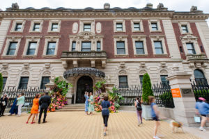 Exterior of the Cooper Hewitt Museum, a three-story red-brick Georgian mansion with elaborate, cream, architectural details, that has eleven people scattered standing in front of it chatting and taking photos