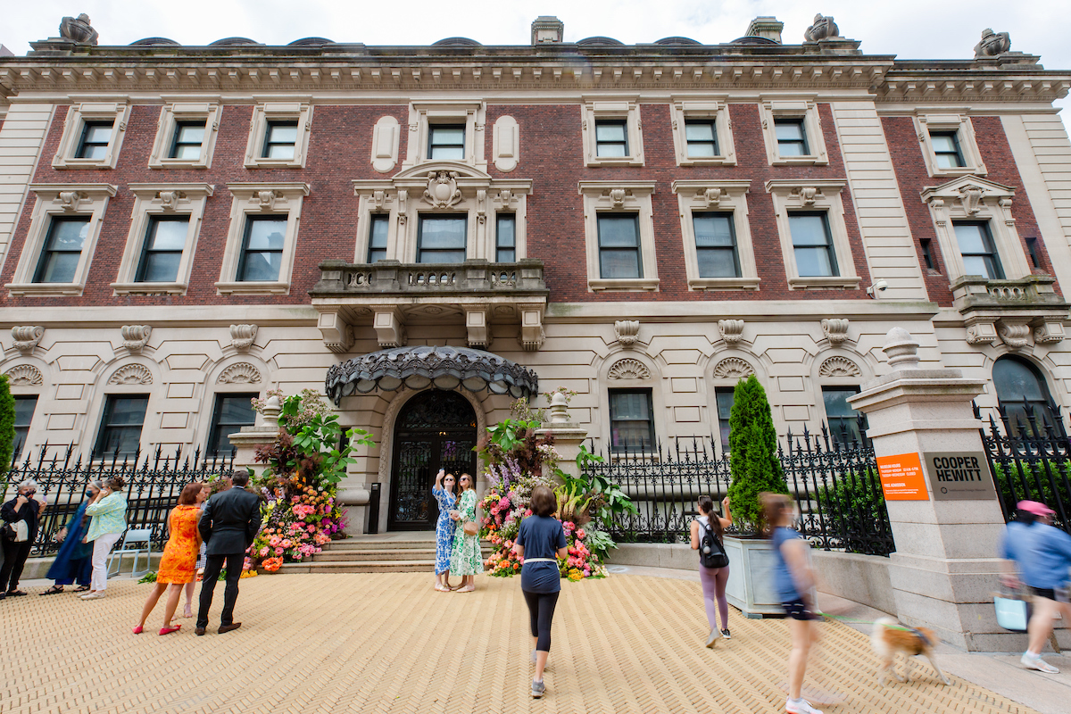Exterior of the Cooper Hewitt Museum, a three-story red-brick Georgian mansion with elaborate, cream, architectural details, that has eleven people scattered standing in front of it chatting and taking photos