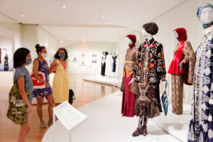 A group of three people stand in an exhibition space discussing four mannequins placed on a raised white stage and dressed in brightly-colored and elaborately patterned dresses, jackets and headscarves