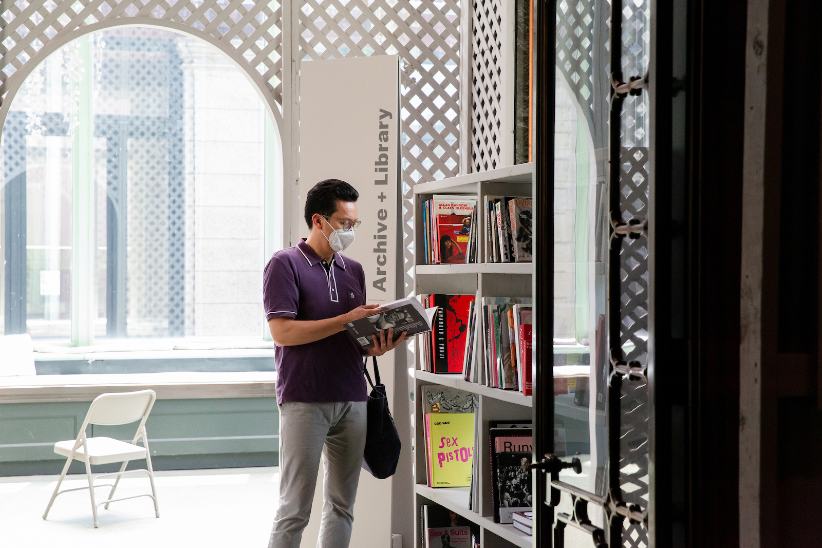 A medium-skinned man with short dark hair and glasses dressed in a purple t-shirt and grey trousers stands in a brightly lit conservatory next to a four-tiered filled book shelf, an open book in his hands