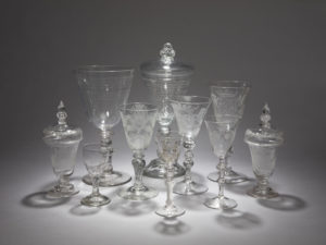 Ten highly decorative eighteenth-century glass vessels arranged in a cluster, including small port glasses, long-stem wine glasses, squat covered vessels, likely for decanting, and tall goblets, photographed against a grey background