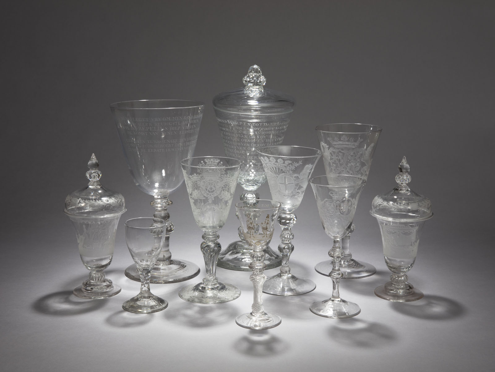 Ten highly decorative eighteenth-century glass vessels arranged in a cluster, including small port glasses, long-stem wine glasses, squat covered vessels, likely for decanting, and tall goblets, photographed against a grey background