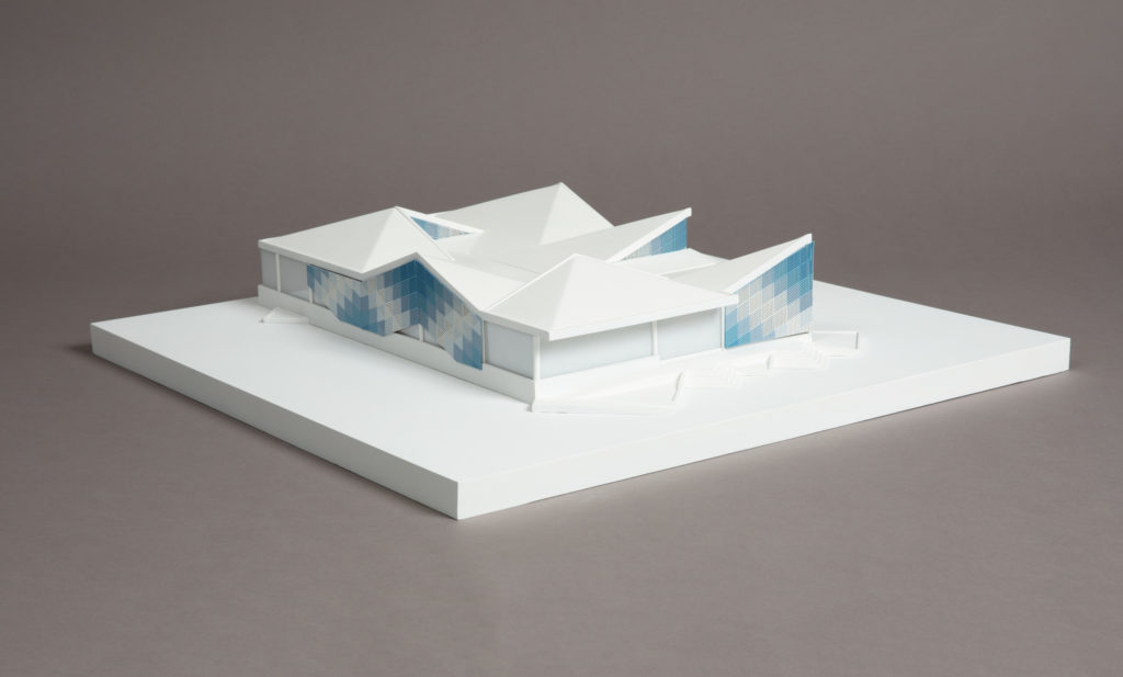 On a gray background, a white three-dimension architectural model of a wide low building with zigzagging roofline and panels adorning the facade in shades of checkered blue