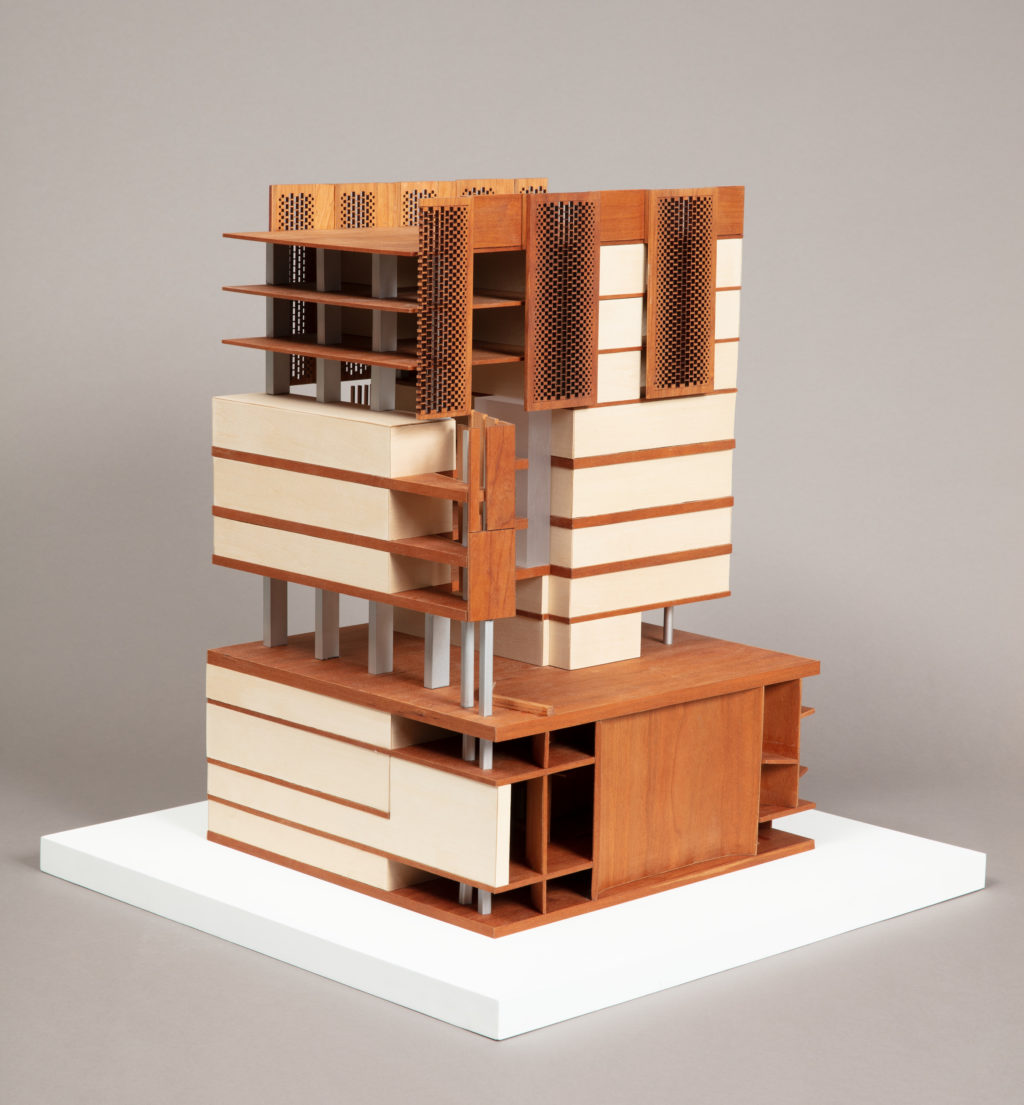 On a gray background, a tall rectangular architectural model in off-white and golden wood tones with long perforated vertical rectangular panels at the upper levels.
