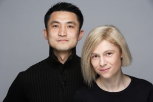 A photograph of a man and woman on a gray background both wearing black.