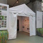 A white pitch-roofed tent installed in the entrance to a windowed conservatory, flanked by hanging white graphic signs and window seats upholstered with shaggy green and multicolor yarn.