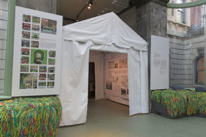 A white pitch-roofed tent installed in the entrance to a windowed conservatory, flanked by hanging white graphic signs and window seats upholstered with shaggy green and multicolor yarn.