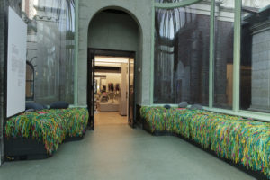 Coolly-lit, green-tiled conservatory, lined with window seats upholstered in shaggy green, yellow, pink, and teal yarns, and scattered with grey pebble-like pillows. Beneath a tall archway at center left, a doorway leads to a warmly-lit shop space.