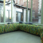 A pale green conservatory space with long bench seating lining its windowed walls, the bench is covered with multi-colored, squiggly strips of recycled fabric in a thick shaggy covering and dotted with grey pebble-like cushions.