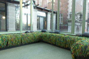 A pale green conservatory space with long bench seating lining its windowed walls, the bench is covered with multi-colored, squiggly strips of recycled fabric in a thick shaggy covering and dotted with grey pebble-like cushions.