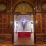 Oak-wood-paneled doorway into a white exhibit space centered in frame, with an installation of a grey standing mannequin, arms at sides, on a plinth behind a transparent screen projected with a person, arms outstretched, overlapping the mannequin.