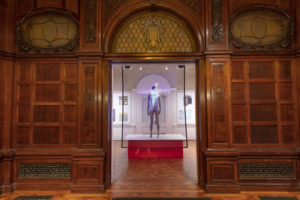 Oak-wood-paneled doorway into a white exhibit space centered in frame, with an installation of a grey standing mannequin, arms at sides, on a plinth behind a transparent screen projected with a person, arms outstretched, overlapping the mannequin.
