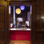 Oak-wood-paneled entryway into an exhibit space centered in frame, a honey-colored armchair with a ribbon-like seat stands side-on on a plinth. Two orb-like yellow and blue lights hang above it, behind is a mannequin wearing blue and white scrubs.