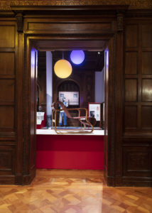 Oak-wood-paneled entryway into an exhibit space centered in frame, a honey-colored armchair with a ribbon-like seat stands side-on on a plinth. Two orb-like yellow and blue lights hang above it, behind is a mannequin wearing blue and white scrubs.