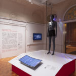 Exhibition space with grey standing mannequin on a plinth in center-left with figural projection on a transparent screen behind, a curved green screen in the foreground and a panel of explanatory text on the white back wall
